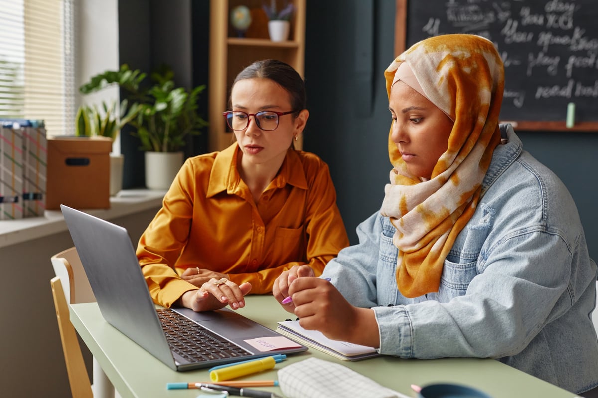 Two young women studying and talking together in a warm classroom — one wearing a hijab — in a supportive, focused conversation